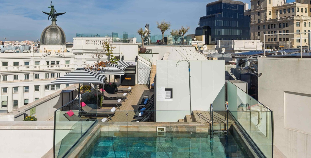 Rooftop plunge pool behind a glass safety rail, with sun loungers on a wooden deck overlooking Madrid's rooftops and a domed building.
