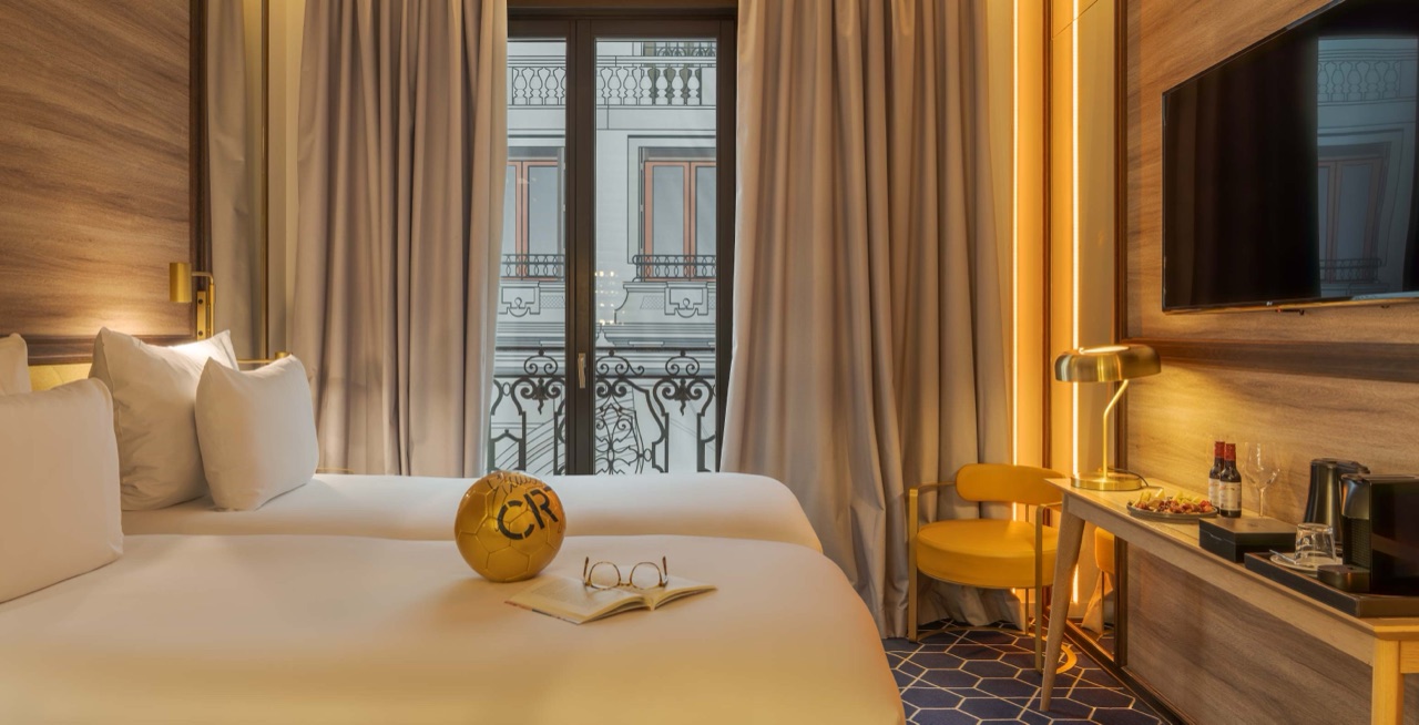Hotel room with a white bed holding a gold branded football and an open book, facing tall curtained French windows with wrought-iron balconies opposite.