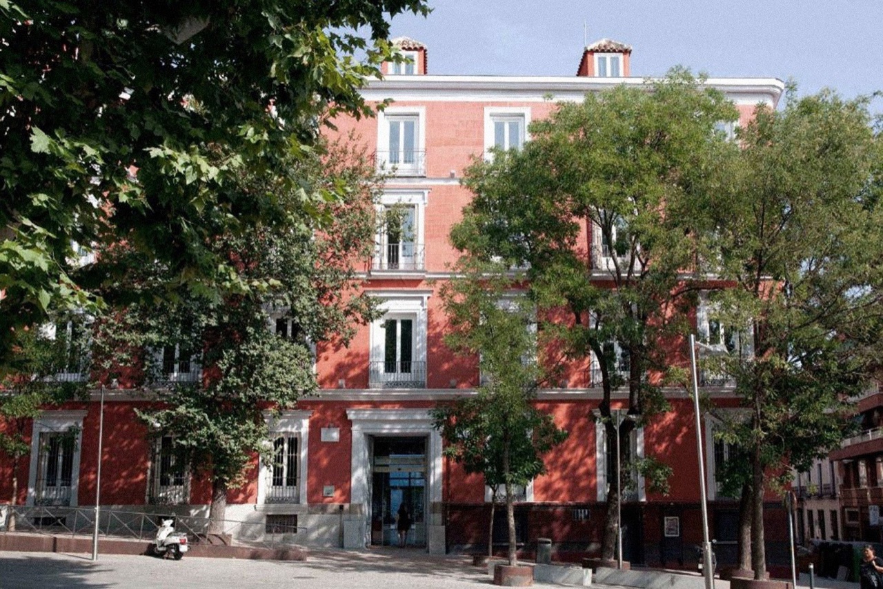 The hotel's red neoclassical facade with white-trimmed balconies and dormer windows, seen from the square through the leaves of street trees.