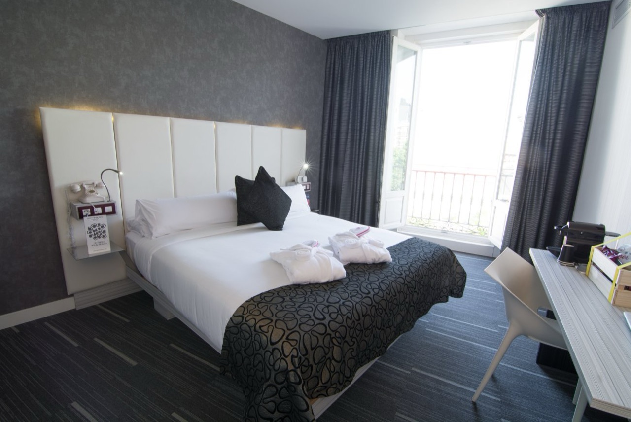 A hotel room with a white bed dressed with folded robes, a dark patterned throw, a padded cream headboard, and open glass doors onto a small balcony.