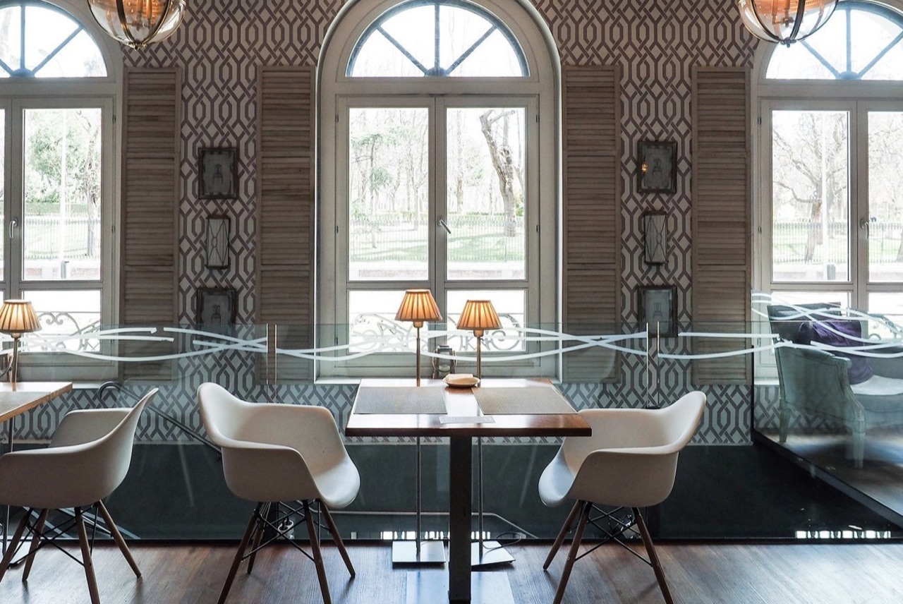 A dining room with tall arched windows looking onto Retiro Park's trees, white shutters and pendant lamps over each table.