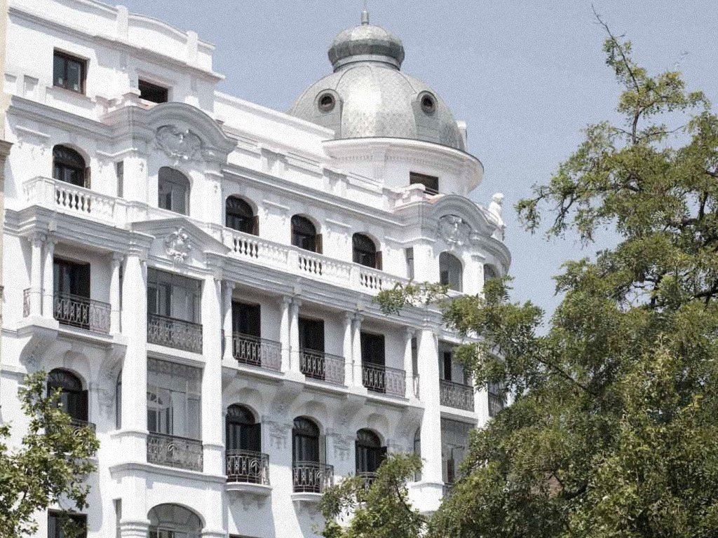 The white facade of Petit Palace Savoy Alfonso XII, with its ornamented balconies and a domed corner tower against a blue sky.