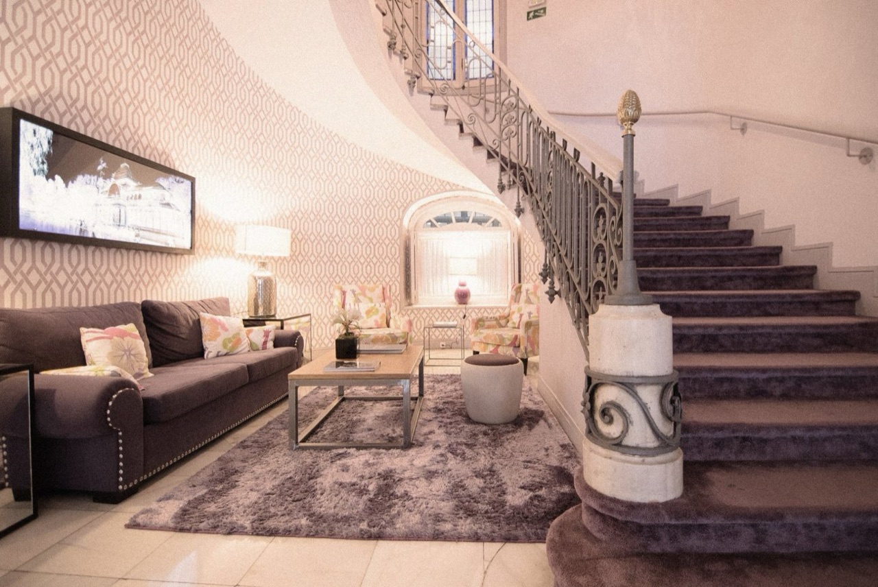 A hotel lobby with a curved wrought-iron staircase, a patterned wallpaper wall, a brown sofa and armchairs with floral cushions.