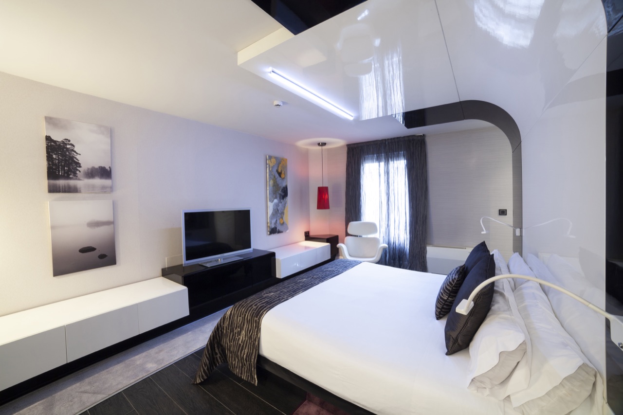 A design-forward guest room with a glossy white ceiling, black accent wall, a red pendant lamp and a white bed.