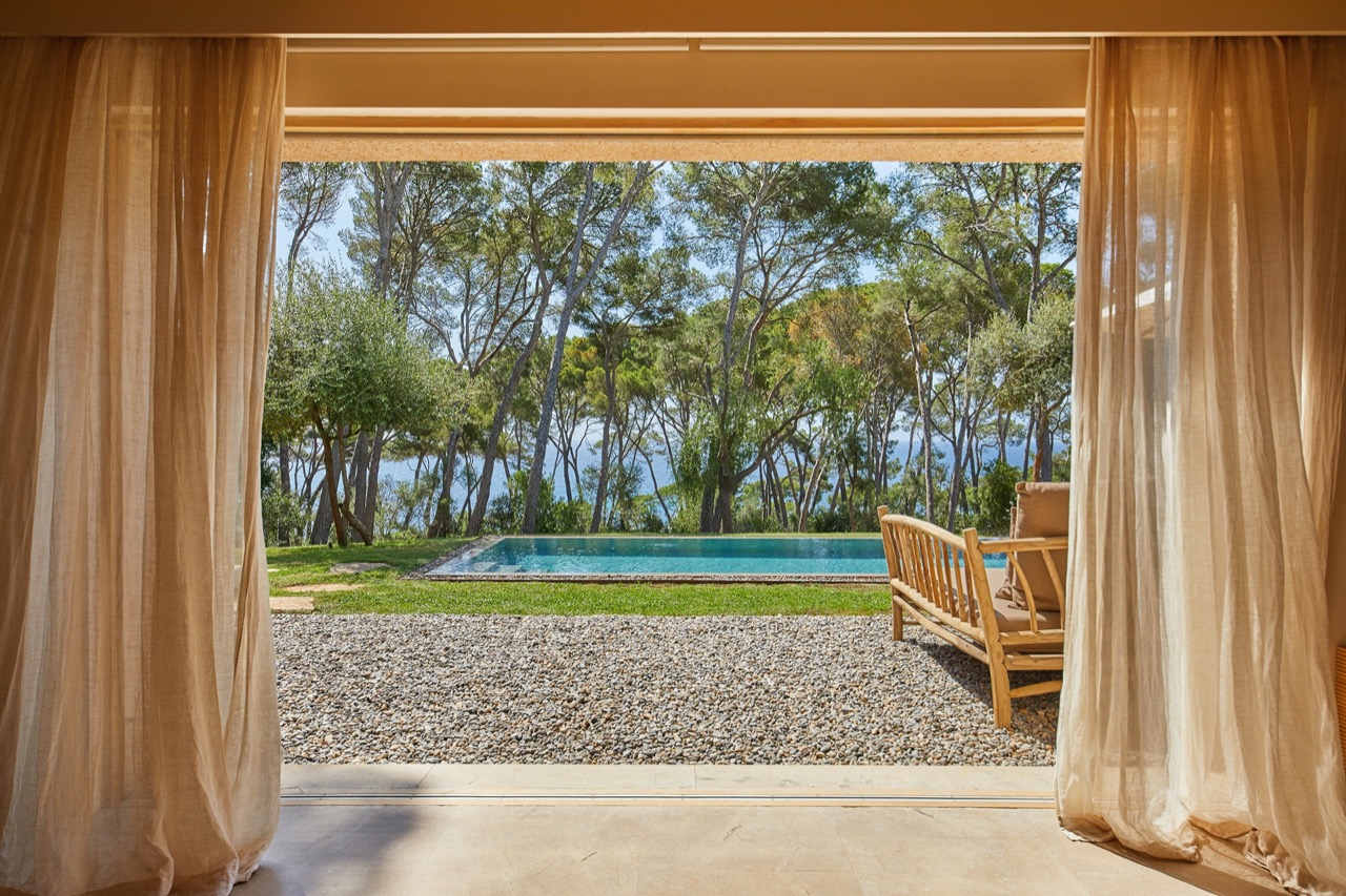View from a suite through open curtains to a pool and pine trees toward the sea