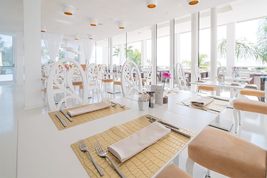 All-white sunlit restaurant at Portals Hills with ornate white chairs, gold placemats and floor-to-ceiling windows onto palm trees