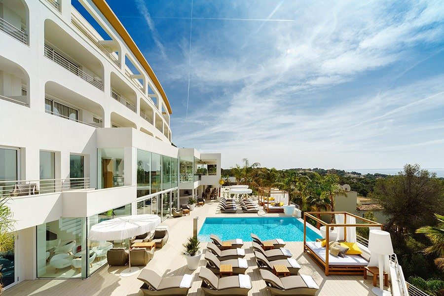 Curved white multi-storey facade of Portals Hills Hotel above its infinity pool, sunbeds and sea view