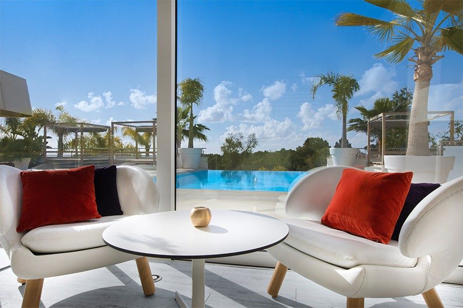 Lobby seating at Portals Hills with white curved chairs and red velvet cushions facing floor-to-ceiling glass onto the infinity pool and palm trees