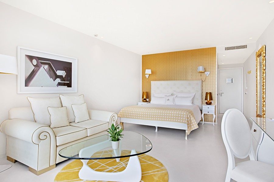 Deluxe suite at Portals Hills with a white tufted headboard against gold patterned wallpaper, a cream sofa and a glass coffee table on a yellow round rug