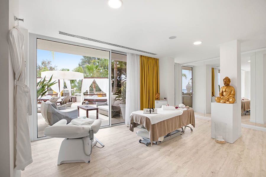 Spa treatment room at Portals Hills with a massage table, a pedicure chair, mustard curtains and a gold Buddha statue, terrace and pool visible through the glass doors