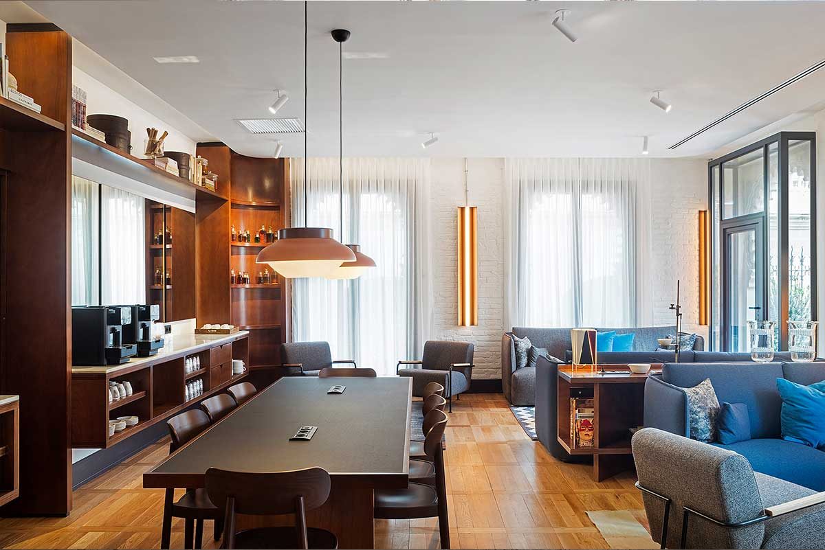 Essens lounge at Praktik Rambla with a dark wood shelving wall, a long communal dining table, orange tube pendant lights, and a blue sofa