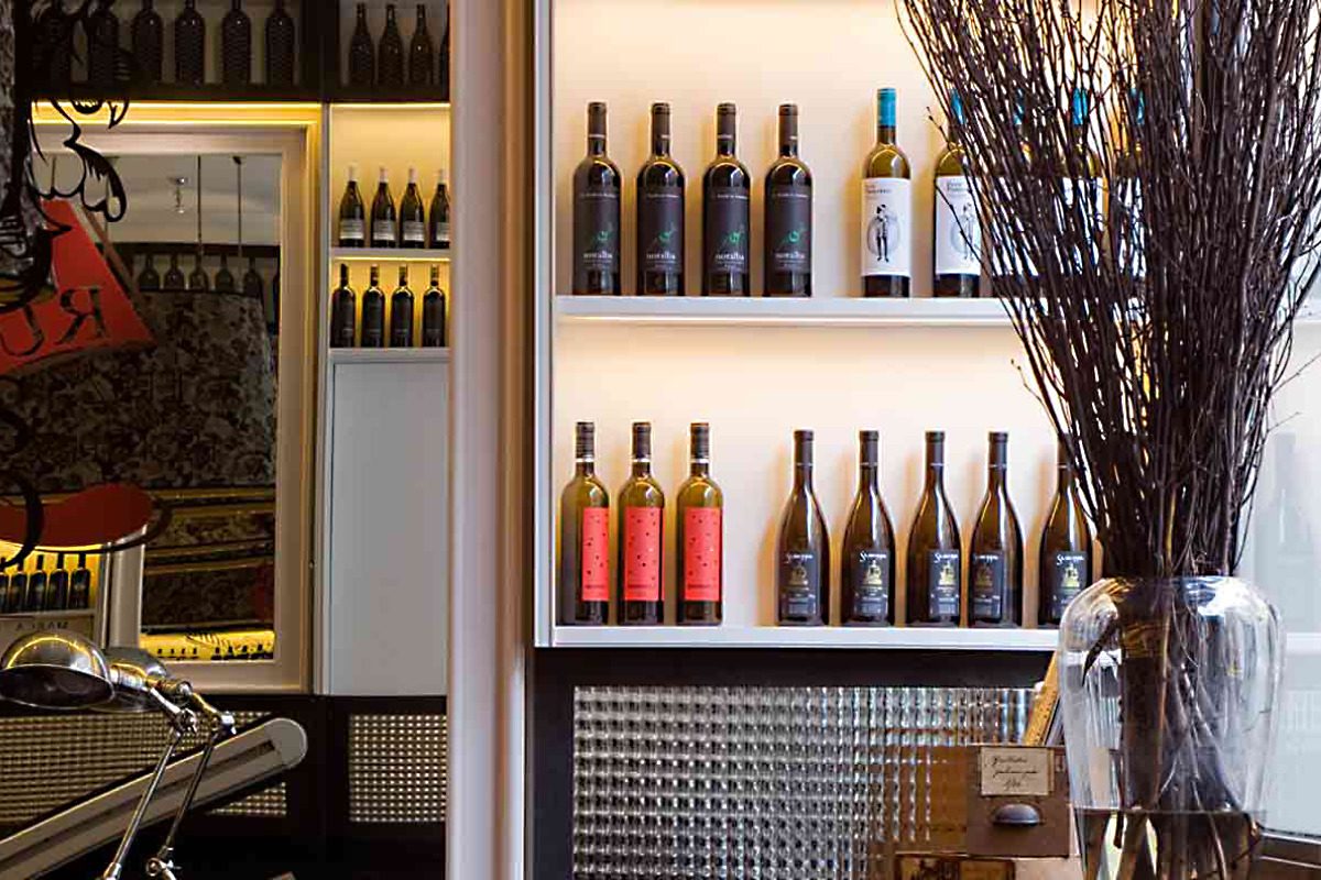 Wine display at Praktik Rambla's vinoteca with backlit shelves of bottles and dried branches in a glass vase