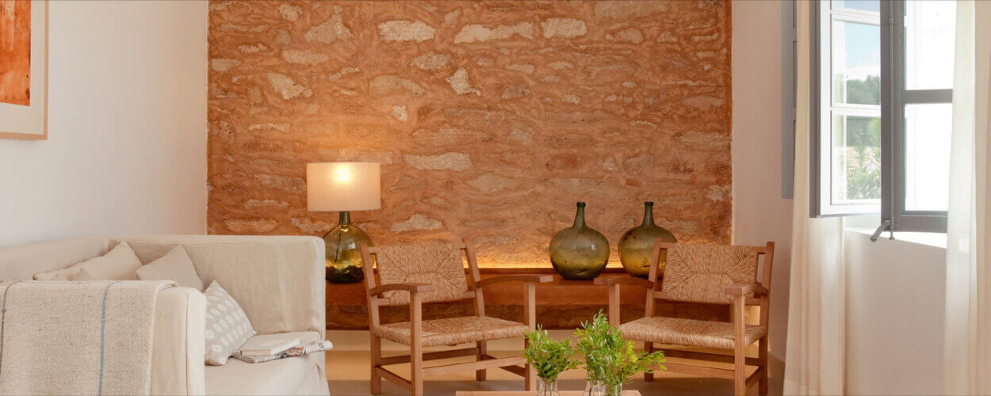 Reading room at Predi Son Jaumell with an exposed sandstone wall, glass demijohn bottles as decor, a cream linen sofa and woven wicker armchairs