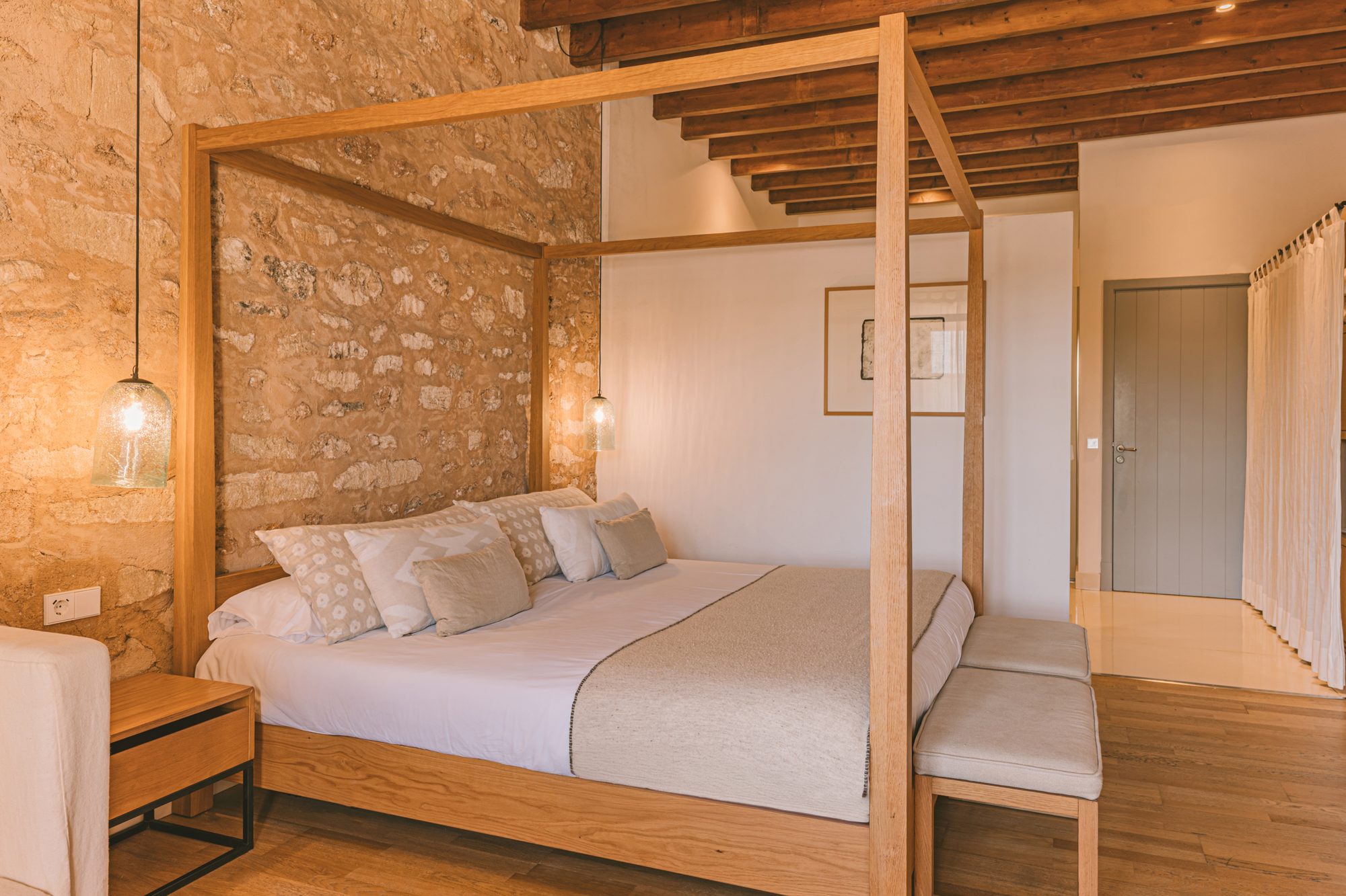 Junior Suite at Predi Son Jaumell with a light wood four-poster bed against an exposed stone wall, under a wood beam ceiling with hanging glass pendant lamps