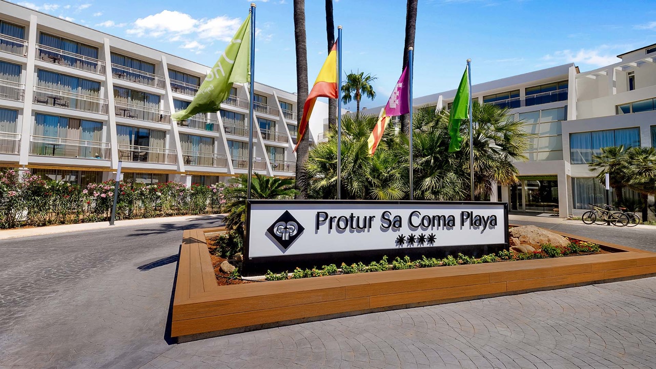 Entrance sign reading Protur Sa Coma Playa with four stars, palm trees and flags in front of the hotel's angular white facade.