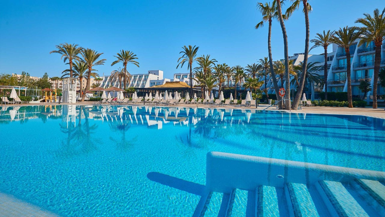 Large outdoor pool at Protur Sa Coma Playa Hotel & Spa lined with sun loungers and white umbrellas under palm trees, hotel buildings behind.