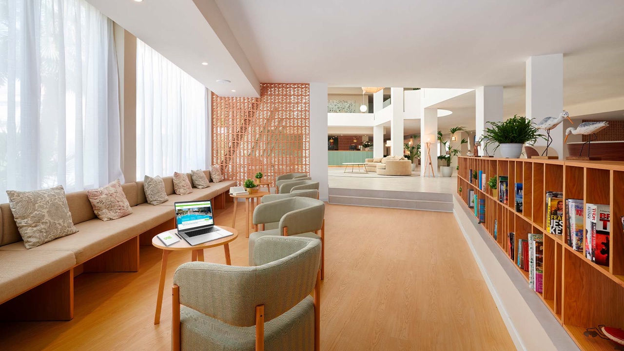 Bright hotel lobby at Protur Sa Coma Playa Hotel & Spa with a built-in bookshelf, lounge seating and the reception desk visible beyond.
