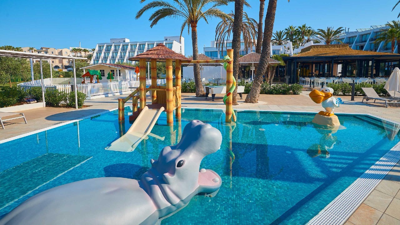 Covered wooden pergola terrace at Protur Sa Coma Playa Hotel & Spa with rattan lounge seating among palm trees.