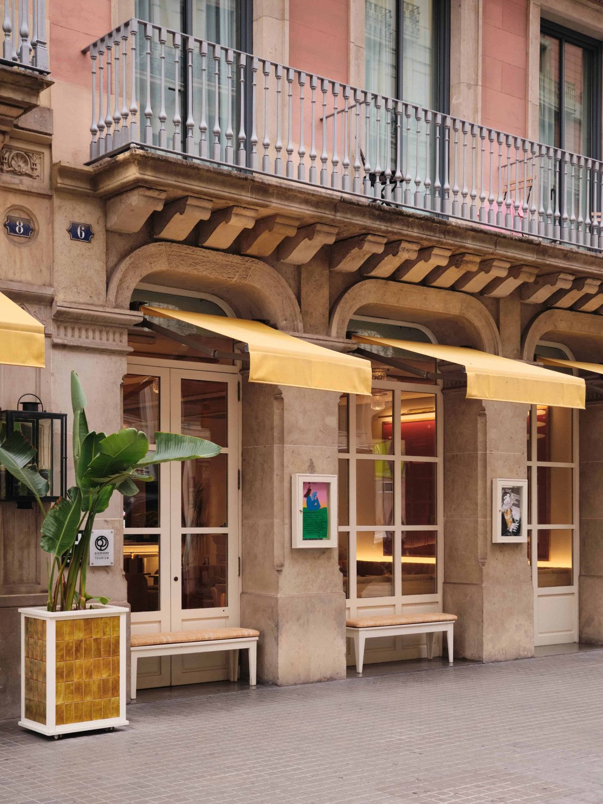 Street facade of Hotel Pulitzer Barcelona with a pink stone building, wrought-iron balconies, and yellow fabric awnings over the ground-floor windows