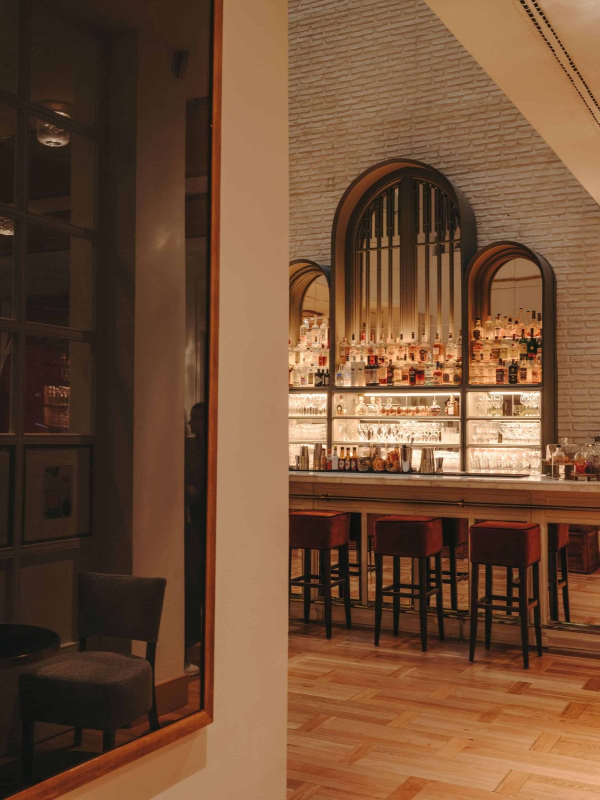 Arched bar at Pulitzer Barcelona with backlit shelves of spirits framed by dark wood arches and red leather stools along the counter