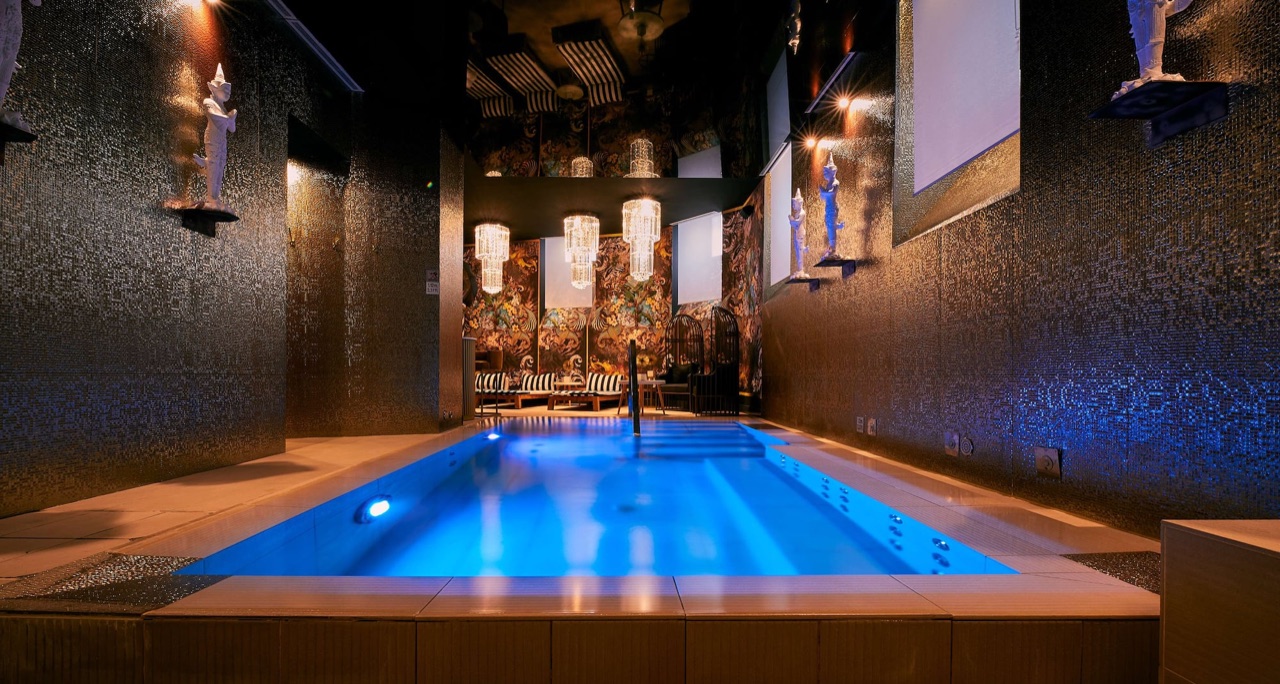The hotel's indoor pool lit blue at night, with mosaic-tiled walls, small statue ornaments on wall shelves, and hanging crystal light fixtures at the far end.