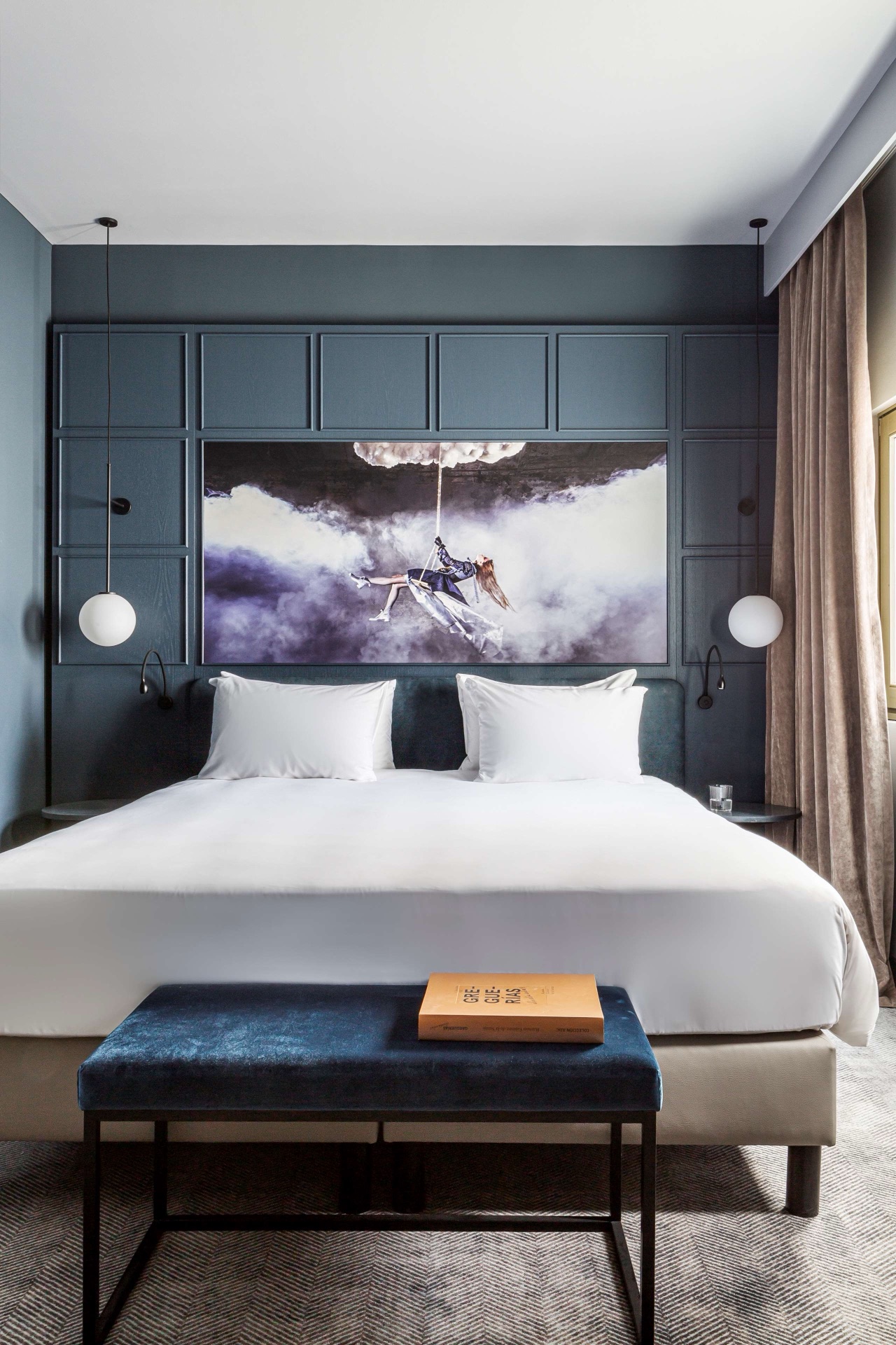 A hotel room with a dark blue panelled headboard, a large framed photograph above the bed, white bedding, and a velvet bench at the foot of the bed.