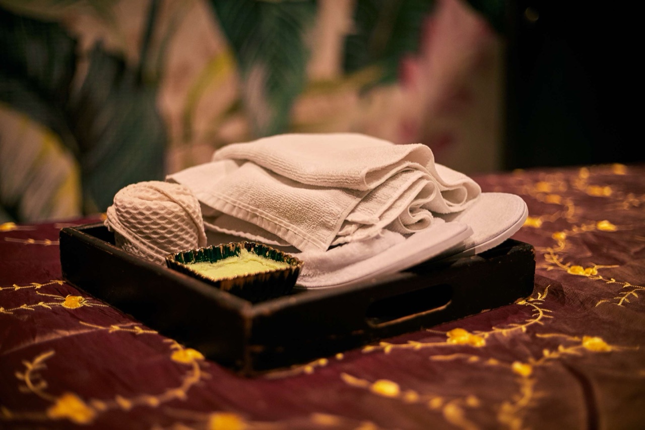 A close-up of folded white spa towels, a loofah, slippers, and a small dish of bath salts on a wooden tray.