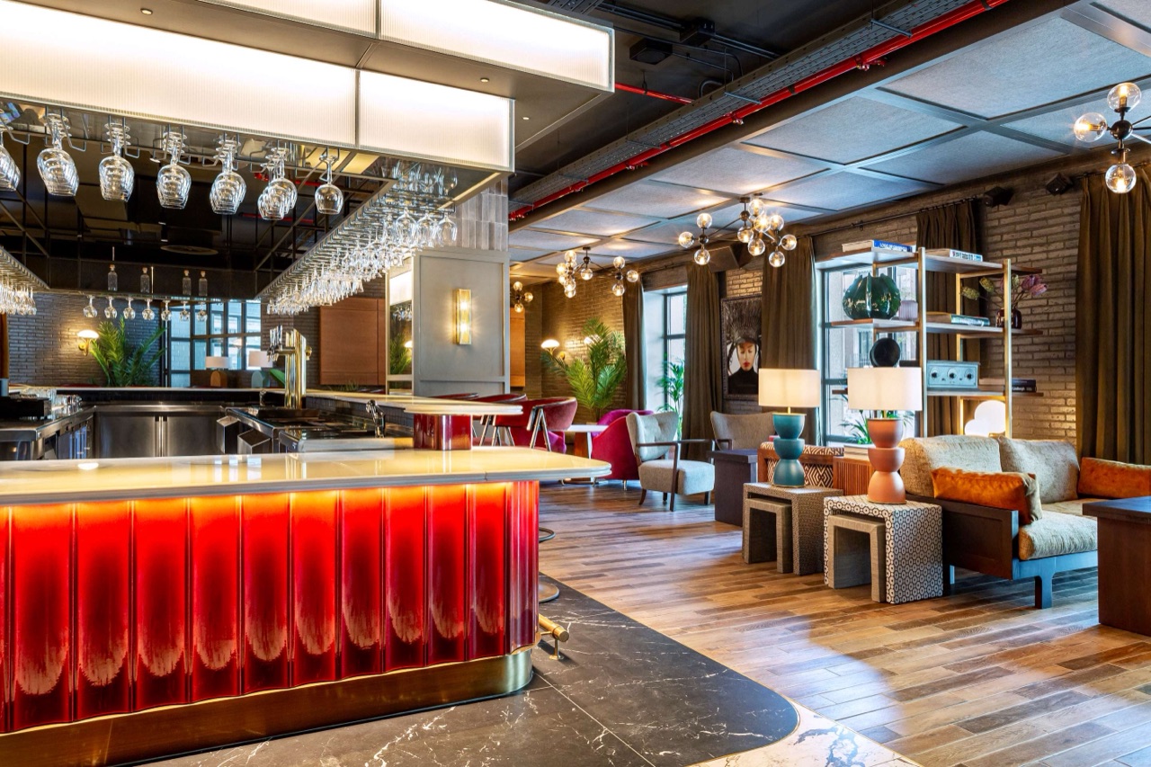 A hotel bar with a glowing red fluted counter, rows of wine glasses hanging above the back bar, and a lounge seating area with armchairs and table lamps beyond.