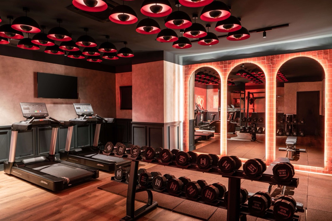 A fitness center with two treadmills, a rack of dumbbells, red dome ceiling lights, and a row of arched, backlit mirrors reflecting more gym equipment.