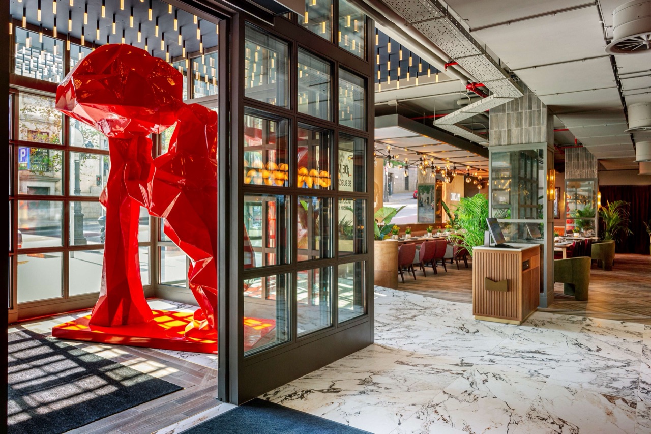 The hotel's glass-fronted entrance with a large faceted red sculpture, opening onto a marble-floored lobby and restaurant seating area beyond.