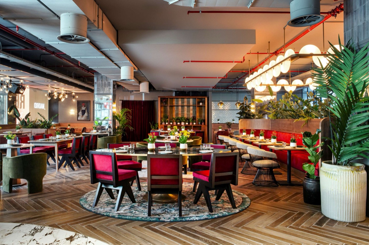 A restaurant dining room with a round table and burgundy velvet chairs on a patterned rug, herringbone wood flooring, a globe-light chandelier, and potted plants throughout.