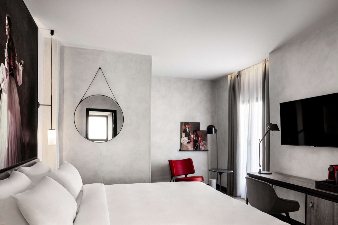 A hotel room with a white bed, a round mirror on a grey wall, a red accent chair, a wall-mounted TV, and a desk with a red coffee machine.