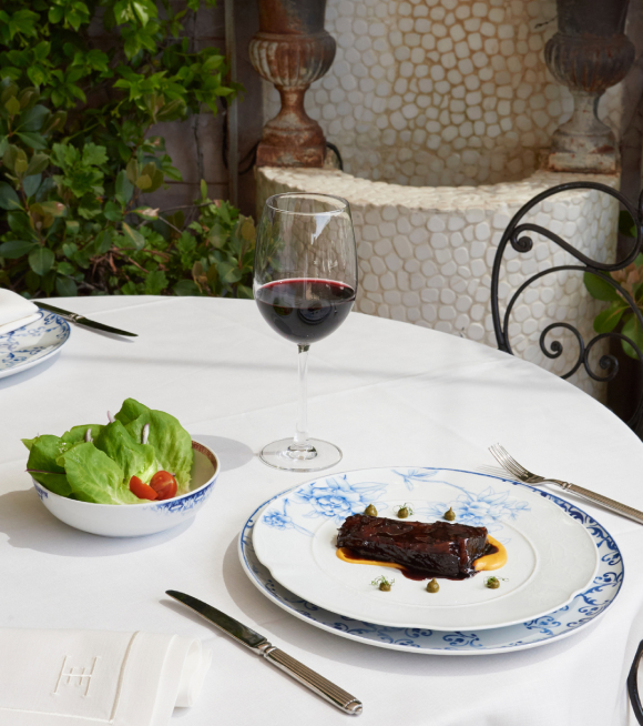 A glazed cut of meat plated on blue-and-white china beside a small salad bowl and a glass of red wine on a white-clothed outdoor table.