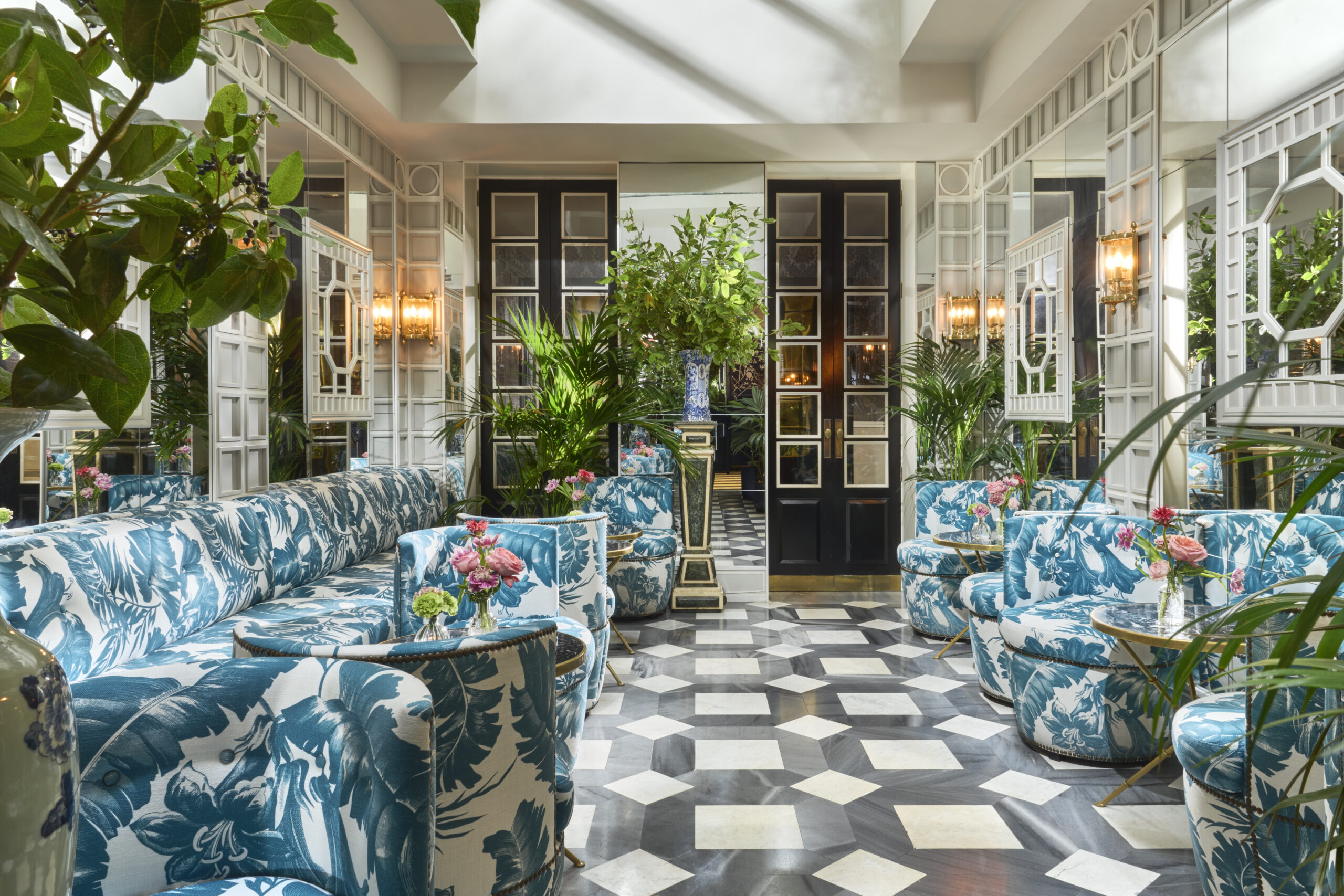 A mirrored salon with blue-and-white palm-print sofas and tub chairs, small marble-topped tables holding vases of pink flowers, potted palms and a diamond-patterned grey-and-white marble floor.
