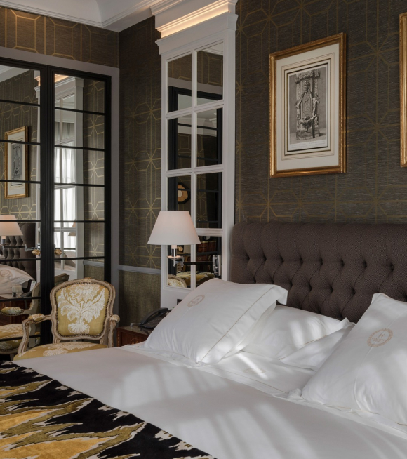 A guest room with a brown tufted headboard, monogrammed white pillows, gilt-framed prints on dark wallpaper and a damask armchair beside a mirrored wardrobe.