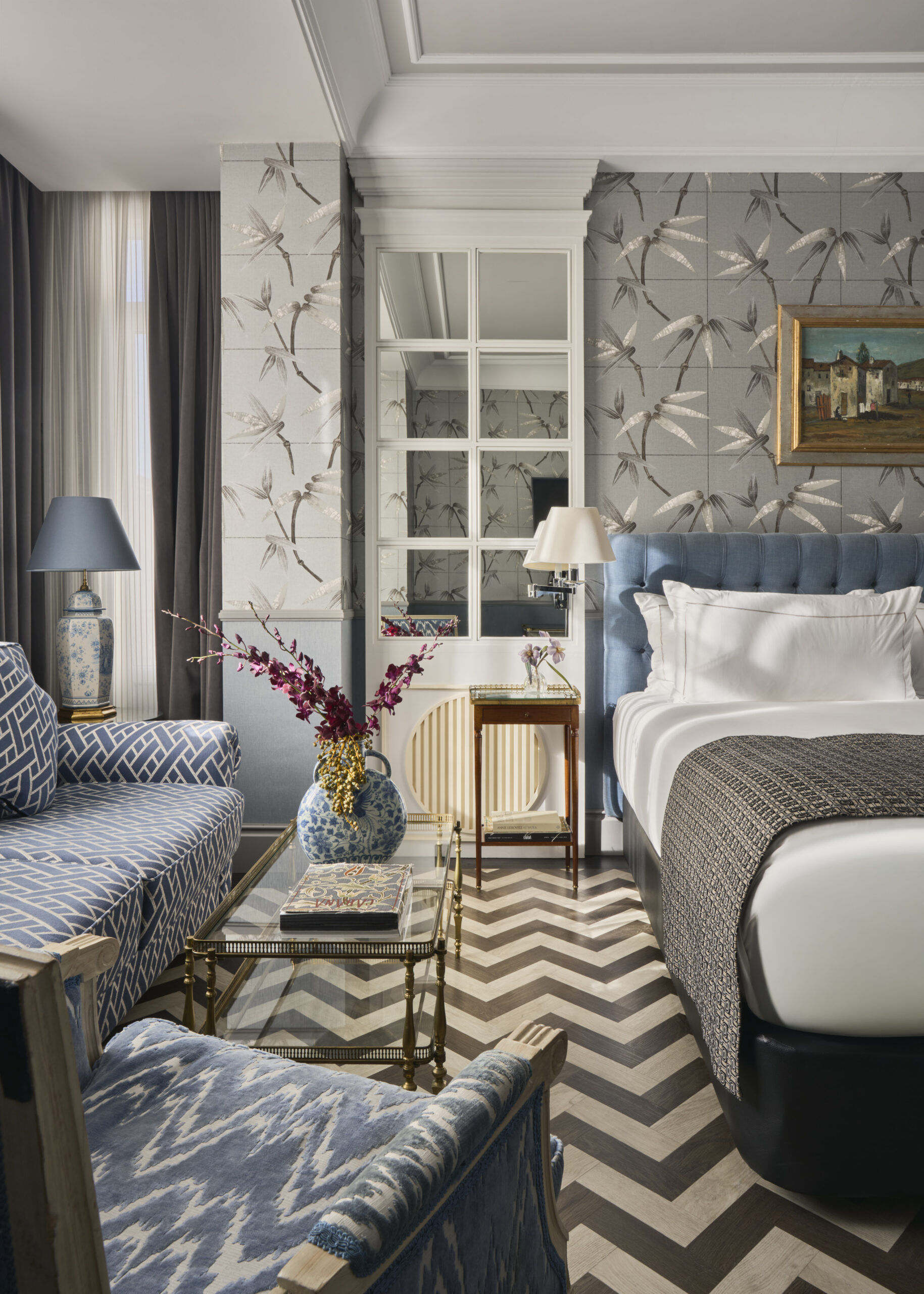 A guest room with a blue tufted headboard, bamboo-print wallpaper, a chevron parquet floor and a blue-and-white patterned seating corner.