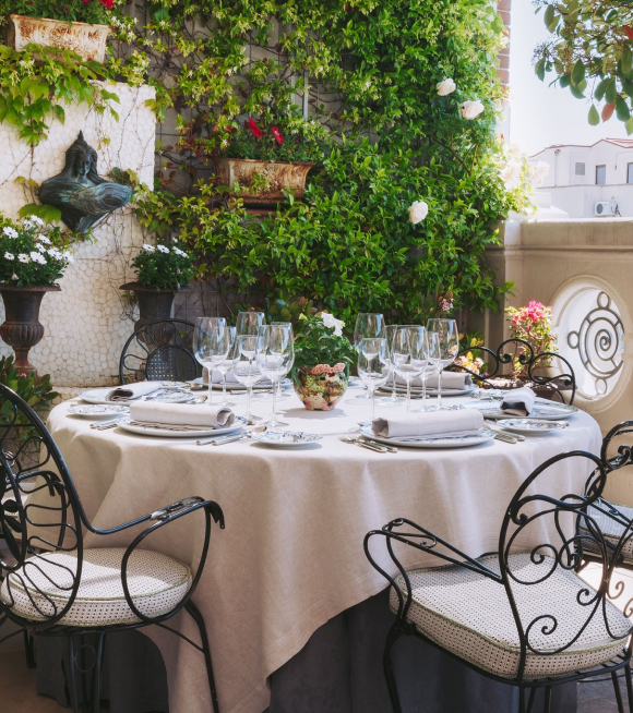 A terrace table dressed in white linen and set with wine glasses and flowers, ringed by wrought-iron chairs against a wall of climbing greenery.