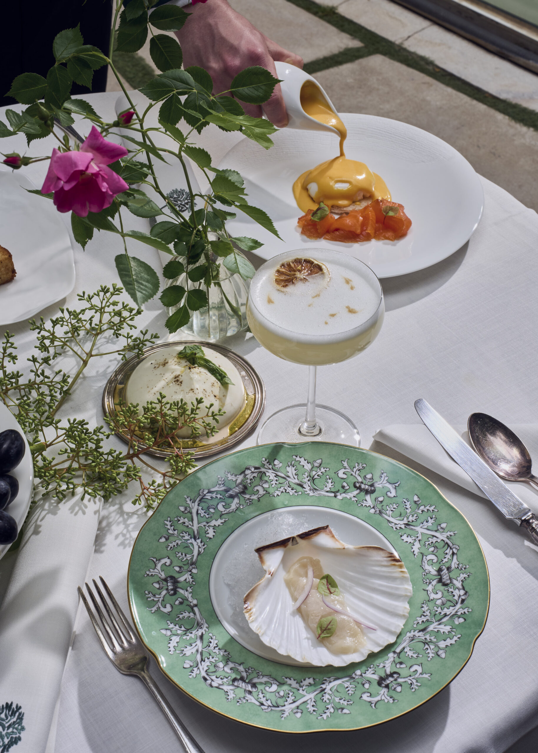 A brunch table with eggs benedict and salmon being sauced from a jug, a foam-topped cocktail, burrata on a silver dish and scallop crudo in its shell on a green-rimmed plate.