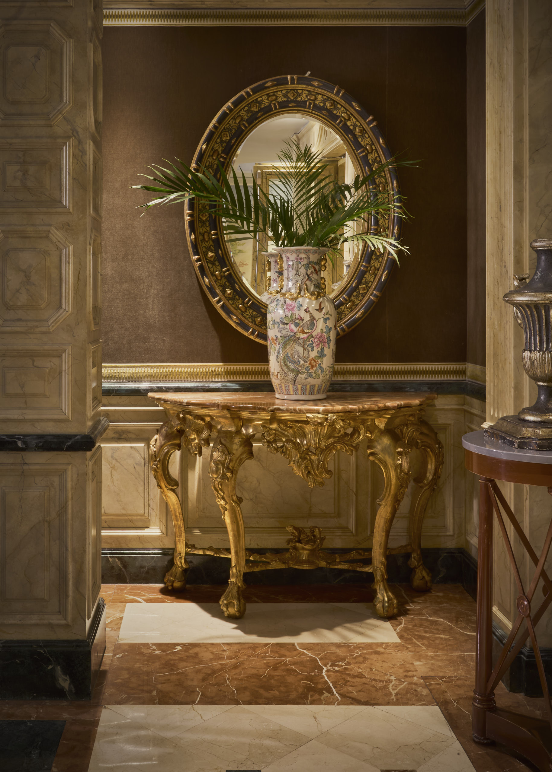 A hallway corner with a gilded marble-topped console, a painted porcelain vase holding palm fronds and an oval gilt mirror against marbled panelling.