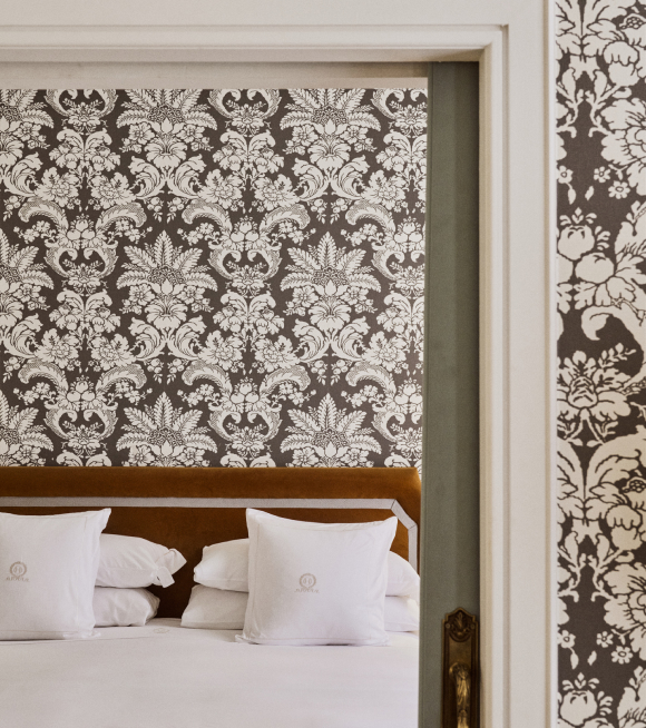A bed with monogrammed white pillows and an amber velvet headboard seen through a doorway, damask-papered walls on either side.