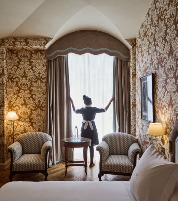 A guest room with brown damask wallpaper, two patterned armchairs and a round table, as a housekeeper draws the curtains at the arched window.