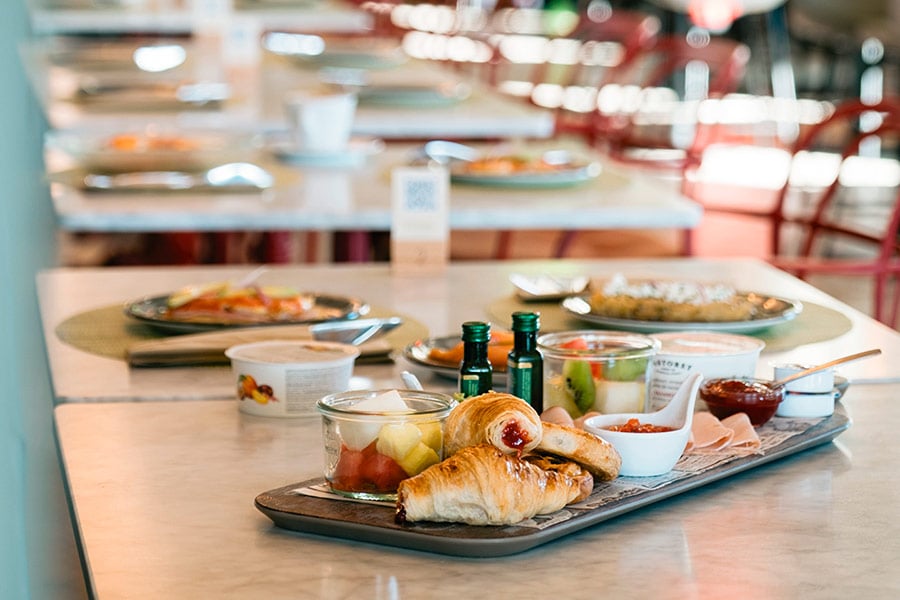 A tray of croissants, cured ham, fresh fruit and small jars of preserves set on a marble table, with rows of tables in soft focus behind it.