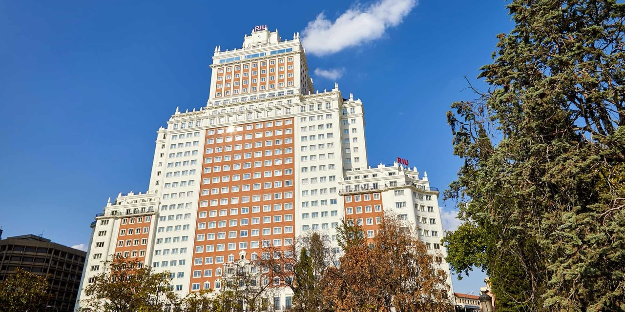 The white and terracotta Edificio España tower rising against a blue sky, with the red RIU sign at its crown, seen from street level through trees.