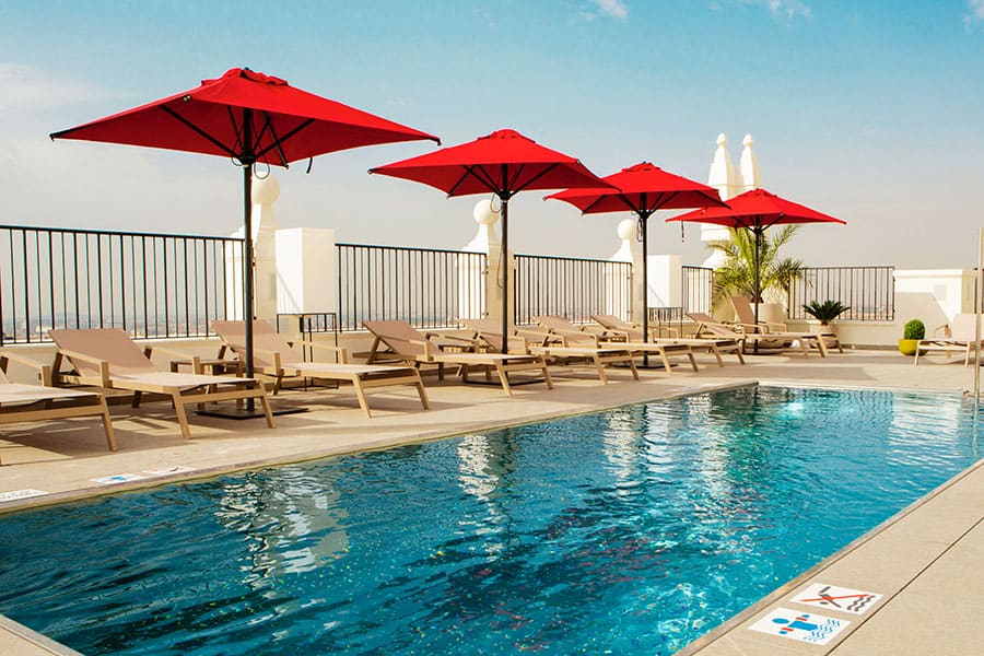 An outdoor rooftop swimming pool lined with sun loungers and red umbrellas, with decorative white finials and a railing along the terrace edge.