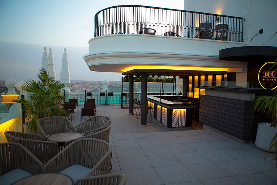 A rooftop terrace bar at dusk with woven armchairs, low tables, and a lit bar counter beneath a round '360°' sign, city rooftops visible beyond the railing.