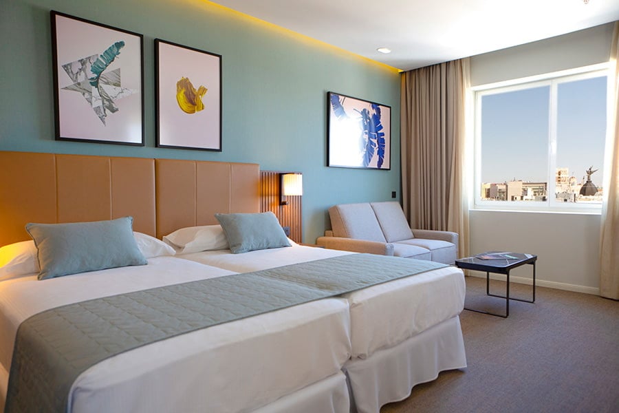 A hotel room with two beds dressed in white and sage-green linen, framed artwork on a teal wall, a sofa, and a window looking out over Madrid rooftops.