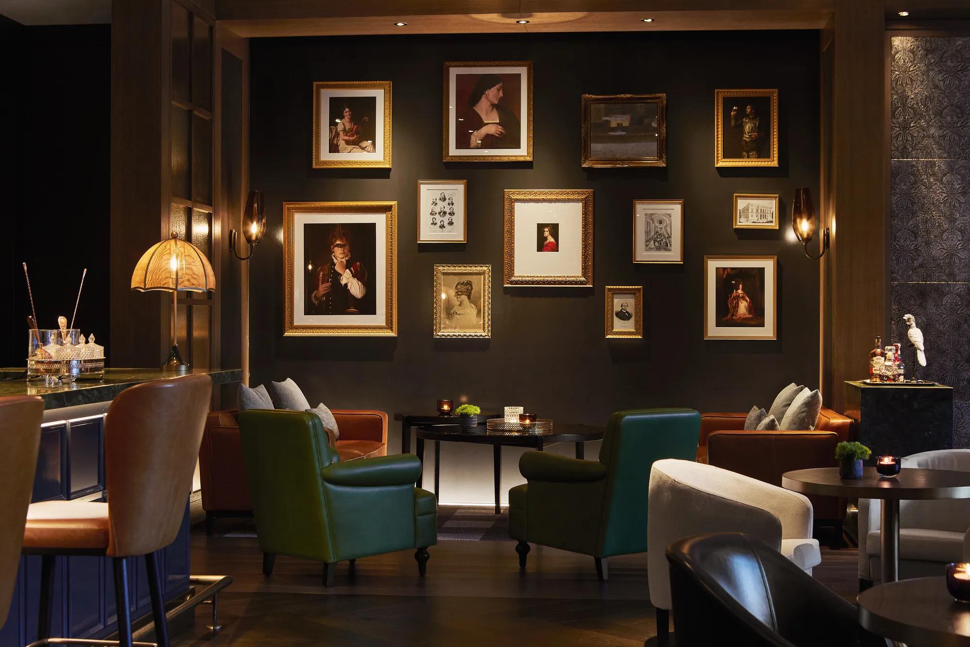 Bar lounge with green and tan leather armchairs, low tables and a wall of gilt-framed portraits under sconce lighting.
