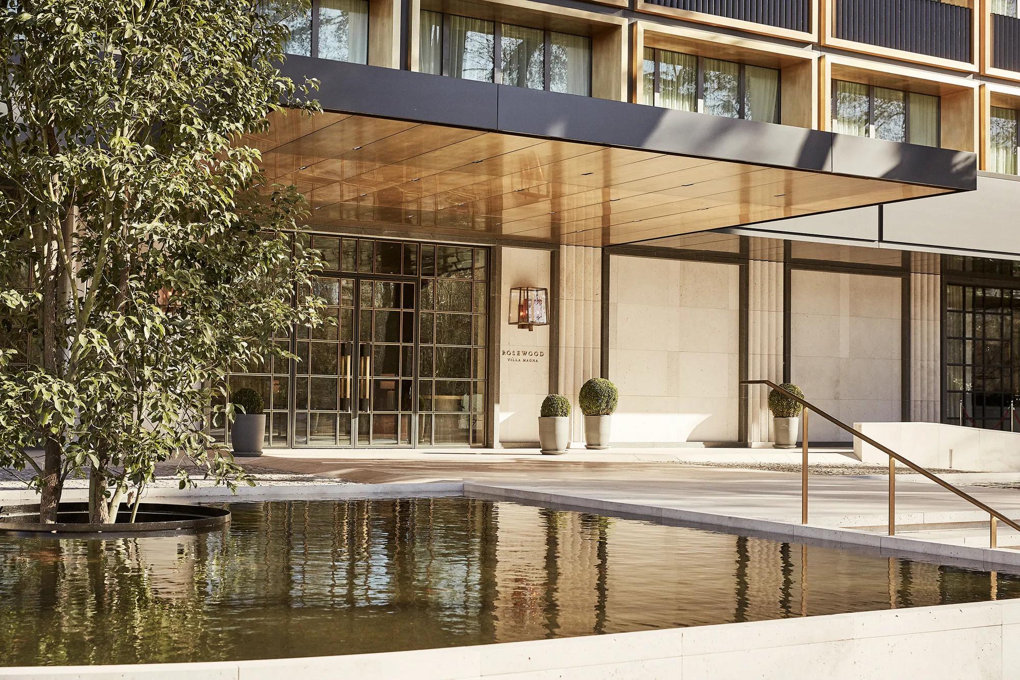 Hotel entrance with a bronze-lined canopy, glass-paned doors, clipped topiary and a reflecting pool, with Rosewood Villa Magna lettering on the stone wall.
