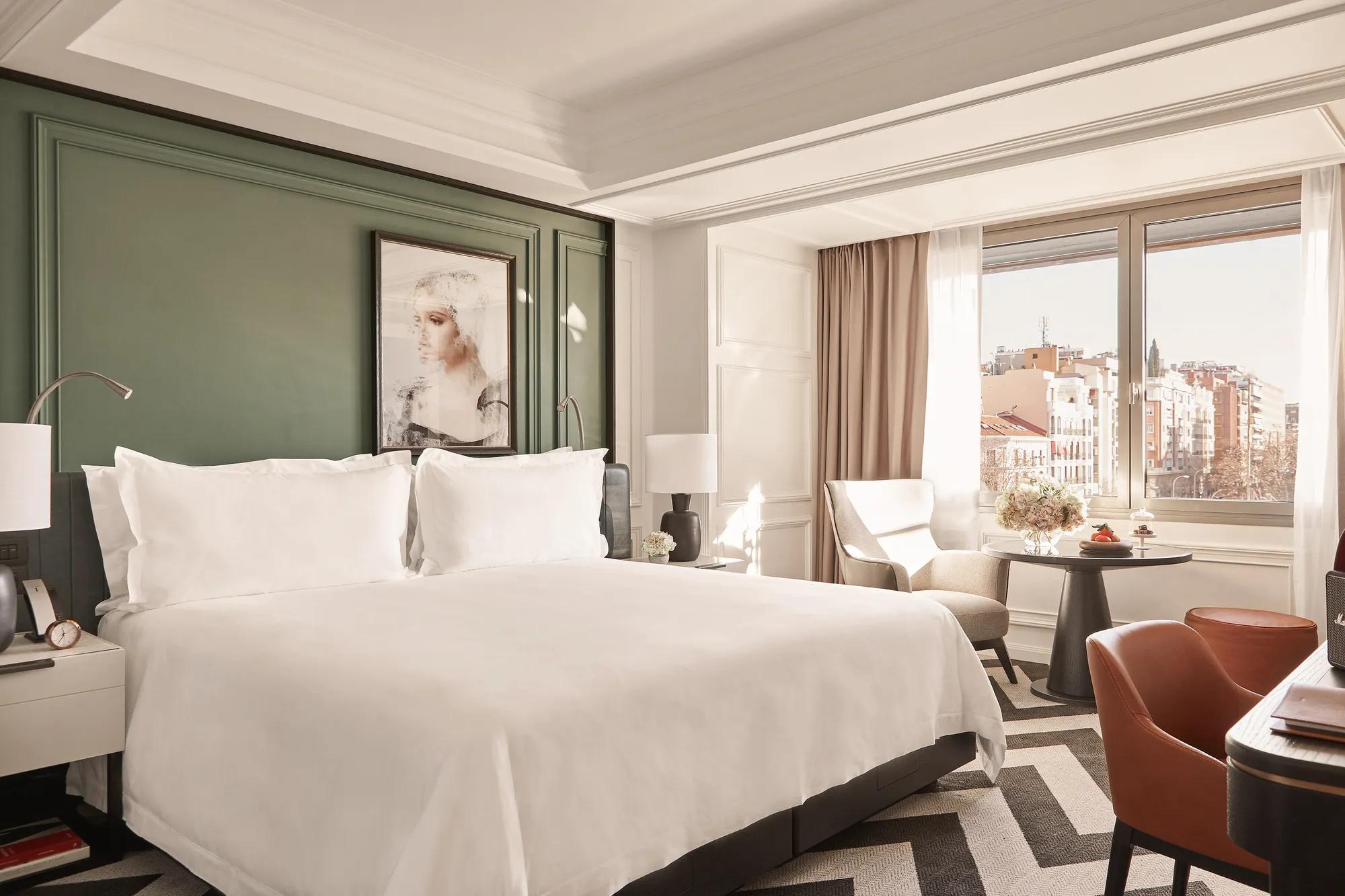 Guest room with a green panelled wall behind the bed, an armchair and round table by the window, and city buildings visible outside.