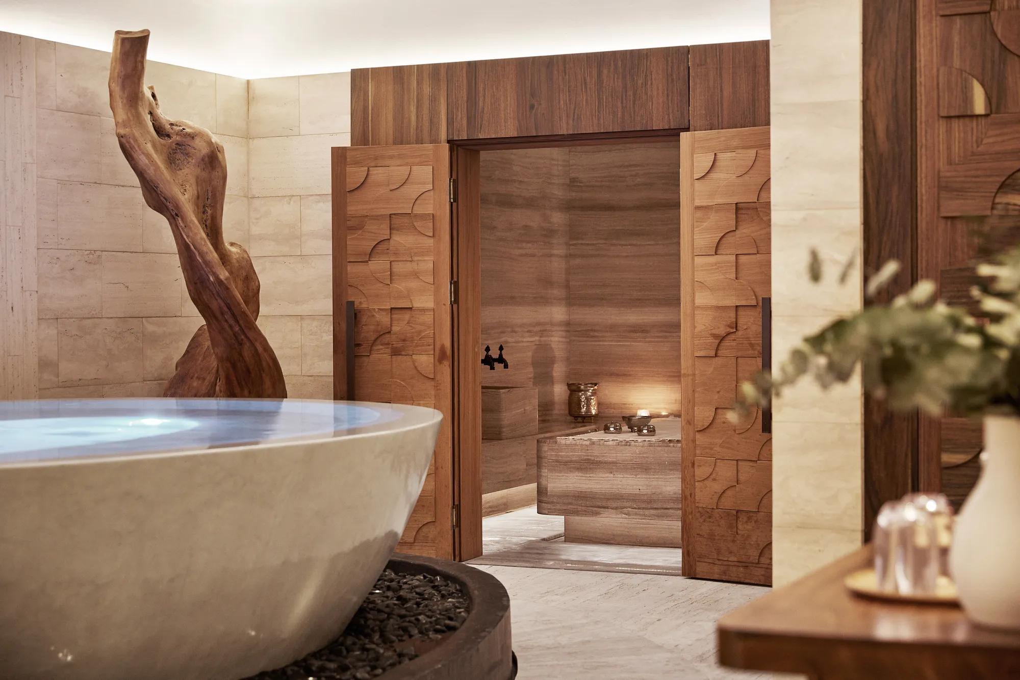 Spa area with a round stone vessel bath, a carved wood sculpture and open panelled doors onto a candlelit stone treatment room.